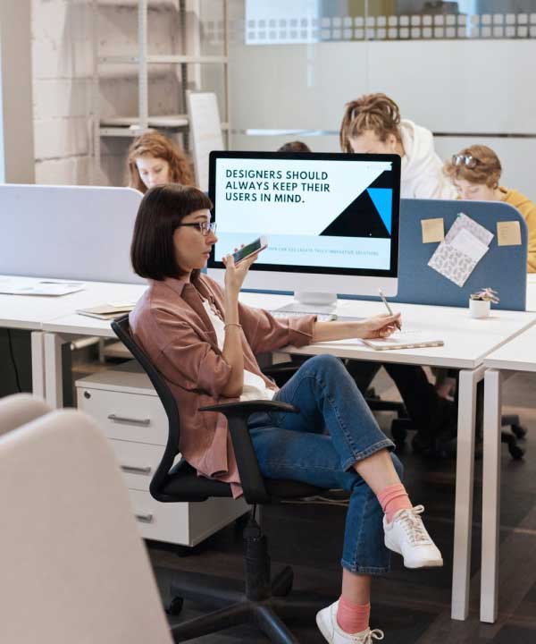 A young woman with short dark hair and glasses sits in a modern office, holding a smartphone and pen, appearing to be in deep thought. She is seated casually in a chair, with one leg crossed over the other. Behind her, a large computer screen displays the text 'Designers should always keep their users in mind.' Other people are working in the background at their desks offering Customer Support Outsourcing services