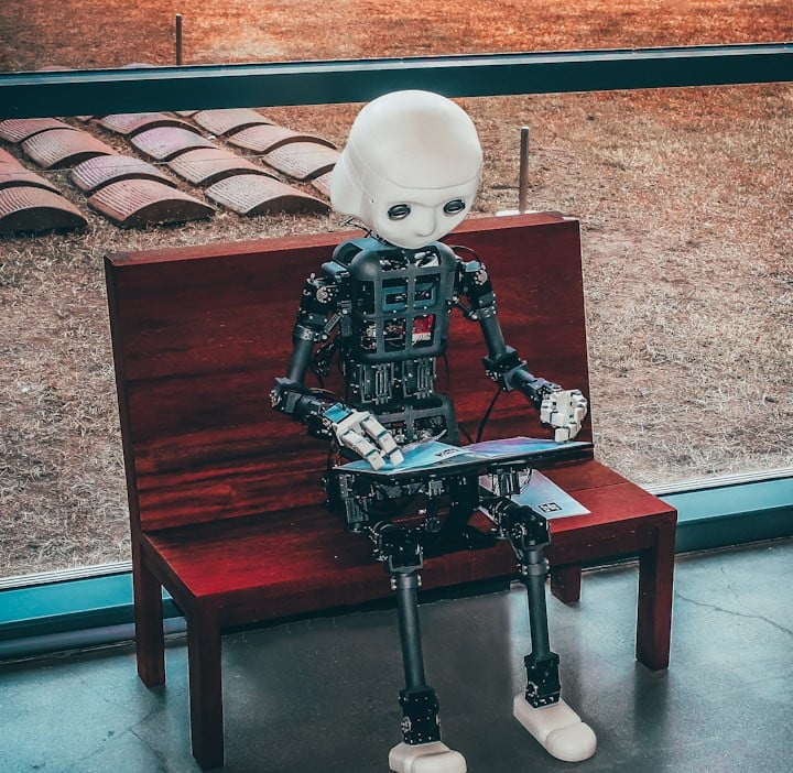 The Role of AI in Modern Customer Support A humanoid robot with a white head and exposed mechanical body parts sits on a wooden bench, interacting with a tablet. The scene is indoors, with a view of the outdoors through a large window in the background. The Role of AI in customer Support Services.